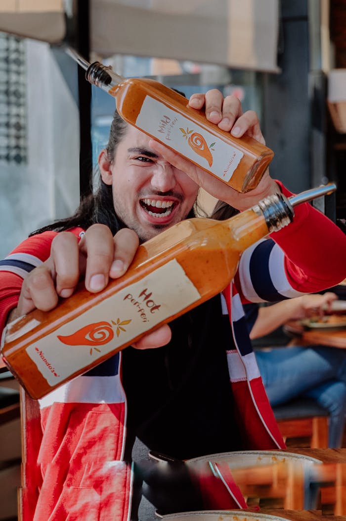 Laughing guy in vivid casual clothes demonstrating bottles with spicy sauce in raised arms while having lunch in modern cafeteria on daytime